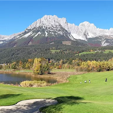 A picturesque golf course landscape with green fairways and a lake. Majestic mountains rise in the background under a clear sky.