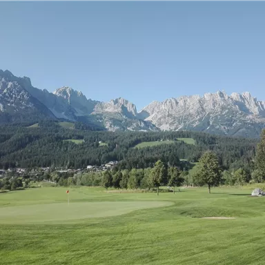 A picturesque golf course landscape with lush green and majestic mountains in the background. The sky is clear and brilliantly blue.