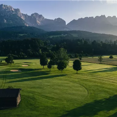 Eine malerische Golfanlage mit gepflegtem Grün und sanften Hügeln. Im Hintergrund ragen majestätische Berge in den Himmel.