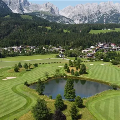 Eine malerische Golfanlage mit gepflegten Rasenflächen und einem kleinen Teich. Im Hintergrund erstrecken sich majestätische Berge und ein charmantes Dorf.