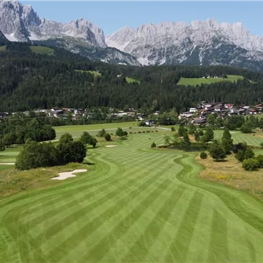 Eine weitläufige Golfanlage mit gepflegten Fairways und Blick auf beeindruckende Berge. Im Hintergrund ist ein kleines Dorf inmitten der Natur zu sehen.