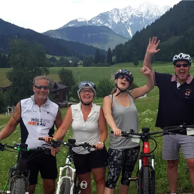 A group of four cyclists is standing happily in a green landscape with mountains in the background. They are posing next to their bicycles and smiling at the camera.