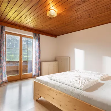 Ein helles Schlafzimmer mit einem Holzdecken und großen Fenstern. Es gibt ein Doppelbett und blumige Vorhänge.
