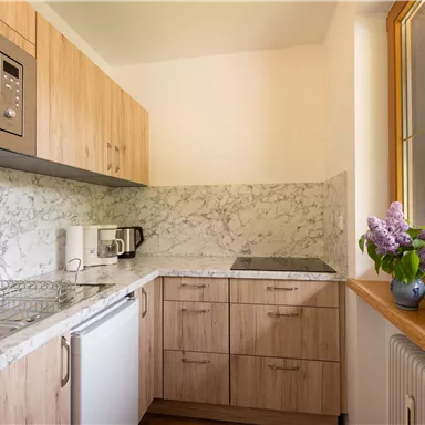 Eine moderne Küche mit Holzmöbeln und einer marmorierten Arbeitsfläche. Das Fenster bietet einen Blick auf die Umgebung und es steht eine Blumenvase mit Flieder auf dem Tisch.