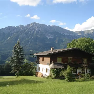 Ein schönes Haus umgeben von grünen Wiesen und hohen Bergen. Der Himmel ist klar und sonnig.
