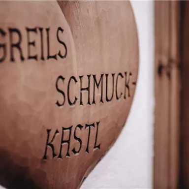 Ein hölzernes Schild mit der Aufschrift „Greils Schmuck-Kastl“. Das Schild hat eine herzförmige Form und zeigt eine rustikale Schrift.