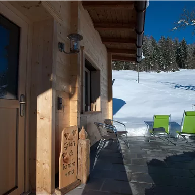 A cozy wooden porch with two green chairs in a snow-covered landscape. The sky is clear and blue, surrounded by trees.