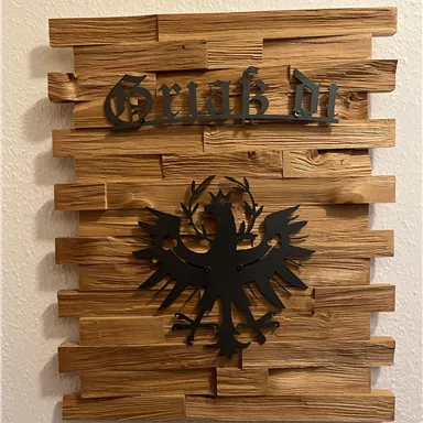 Ein dekoratives Holzschild mit dem Schriftzug "Grüß dich" und einem schwarzen Wappen. Das Design kombiniert rustikale Holzlatten mit einem auffälligen Symbol.