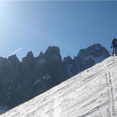 Zwei Personen steigen einen schneebedeckten Hang hinauf. Im Hintergrund sind hohe Berge und ein klarer blauer Himmel zu sehen.