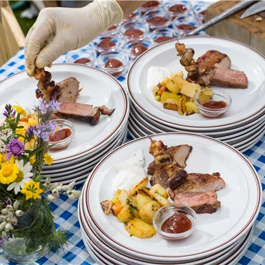 Ein Tisch mit mehreren Tellern, die gegrilltes Fleisch und Beilagen wie Kartoffeln und Soße enthalten. Dazu stehen Blumen in einer Vase.
