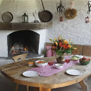 A rustic dining area with a large wooden table and a fireplace. The table is set with various dishes and fresh flowers in a vase.