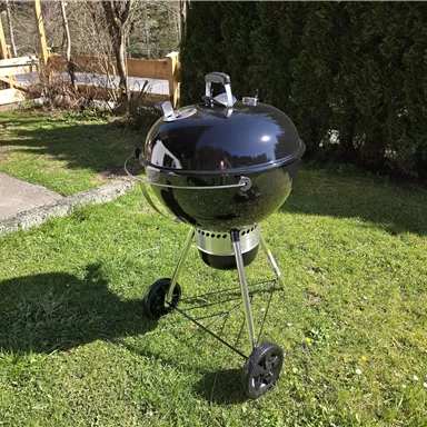 Ein runder Schwarzgrill steht auf grünem Gras. Im Hintergrund sind Bäume und eine Terrasse zu sehen.