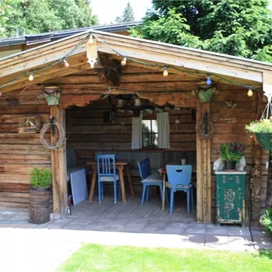 Eine gemütliche Holzhütte mit einem rustikalen Charme. Der Außenbereich ist mit Pflanzen und bunten Lichtern dekoriert.