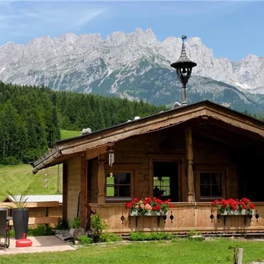 Eine gemütliche Holzhütte umgeben von grünen Wiesen und Bergen. Blumen schmücken die Fenster, und die Landschaft ist malerisch.