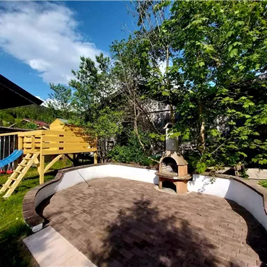 An inviting garden with a stone terrace and a barbecue area. In the background, there is a play tower and there is plenty of green vegetation.