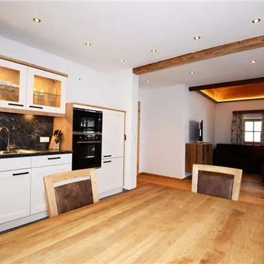 Eine moderne Küche mit einem großen Esstisch aus Holz und hochwertigen Geräten. Im Hintergrund befindet sich ein gemütlicher Wohnbereich mit Sitzgelegenheiten.