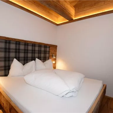 Ein gemütliches Schlafzimmer mit einem Doppelbett und Holzdetails. Die Wände sind schlicht gehalten, und ein Fenster mit einem grünen Vorhang lässt Tageslicht herein.