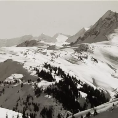 A snow-covered mountain landscape with high peaks and deep valleys. The gentle hills are lined with trees.