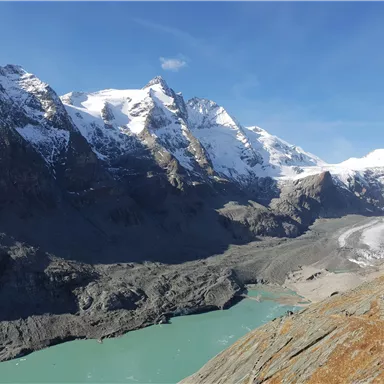 An impressive mountain landscape with snow-covered peaks and a clear turquoise lake. The sky is blue, and the surroundings appear majestic.