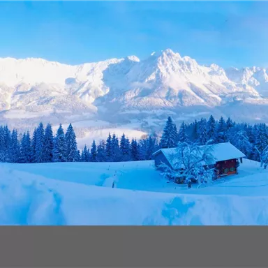 A winter landscape with snow-covered mountains and a small wooden house. The sky is clear and blue, surrounded by snow-covered trees.