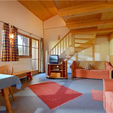 A cozy living room with wooden paneling and stairs. There is a sofa, a table, and large windows that let in plenty of light.