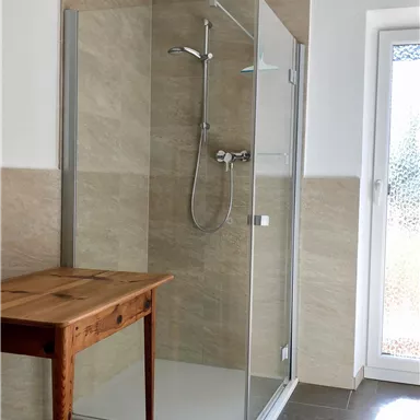 Modernes Badezimmer mit einer klaren Duschkabine und einem Holztisch. Ein Fenster lässt Tageslicht herein.