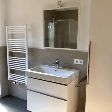 Ein modernes Badezimmer mit einem weißen Waschbecken und einem großen Spiegel. Die Wände sind in neutralen Farben gehalten, und ein Handtuchradiator ist an der Wand montiert.