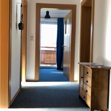 Ein heller Flur mit Holzrahmen und Teppichboden. Am Ende des Flurs ist ein Fenster und eine blau Vorhang.