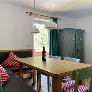 Ein gemütlicher Essbereich mit einem Holztisch und bunten Stühlen. Vor dem Fenster stehen grüne Vorhänge und eine Flasche Wein auf dem Tisch.