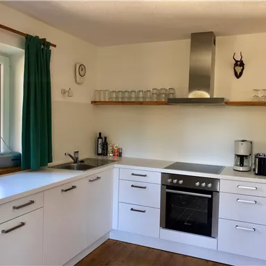 Moderne Küche mit weißen Möbeln und einer Kochstelle. Das Fenster lässt viel Tageslicht herein, und es gibt eine grüne Vorhang.