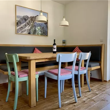 Ein gemütlicher Essbereich mit einem Holztisch und bunten Stühlen. Der Raum hat große Fenster und eine Kose für eine freundliche Atmosphäre.