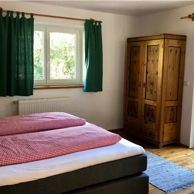Ein gemütliches Schlafzimmer mit einem großen Bett und rot-weiß karierten Bettdecken. An den Fenstern hängen grüne Vorhänge, und ein Holzkasten steht in der Ecke.