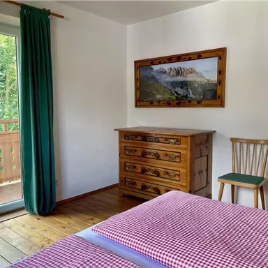 Ein gemütliches Schlafzimmer mit einem Doppelbett und einer Holzkommode. Große Fenster mit Blick auf einen Balkon und grüne Vorhänge sorgen für Licht und Frische.