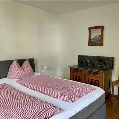 Ein gemütliches Schlafzimmer mit einem großen Doppelbett und karierten Kissen. Neben dem Bett steht ein Holzstuhl und eine Kommode mit einer grünen Marmoroberfläche.