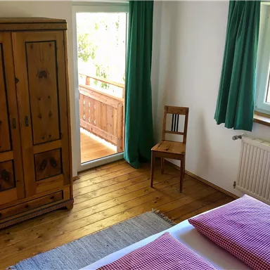 Ein gemütliches Schlafzimmer mit einem Holzschrank und einem Stuhl. Durch das Fenster ist ein schöner Ausblick auf einen Balkon zu sehen.