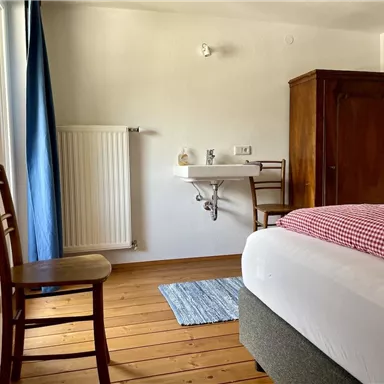 Ein einfaches Zimmer mit einem Bett, einem Holzschrank und einem Waschbecken. Der Raum hat einen Holzfußboden und ein Fenster mit blauen Vorhängen.