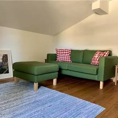 Ein gemütliches, grünes Sofa mit karierten Kissen und einem passenden Hocker. Der Raum hat Holzböden und einen bunten Teppich.