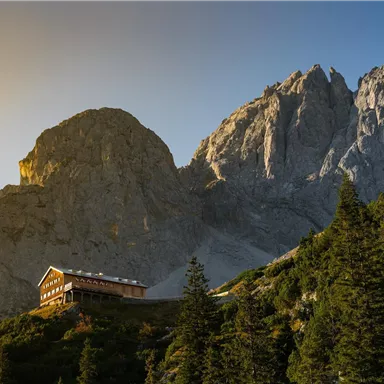 A picturesque mountain panorama with tall rocks and a cottage in the foreground. The sun gently shines on the landscape and the trees.