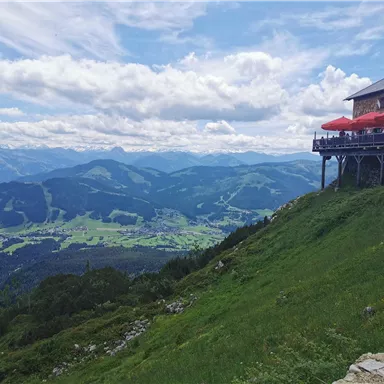 A mountain landscape with a restaurant that has a terrace with red umbrellas. The green meadows and the expansive valley provide a beautiful view of the surrounding mountains.