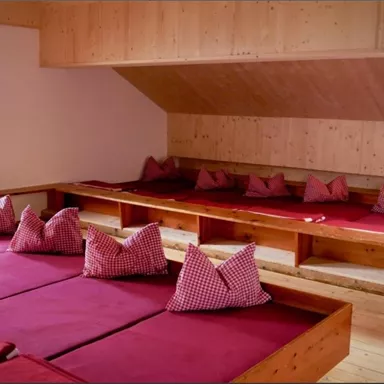 A cozy room with low wooden platforms and red cushions. The walls are paneled with wood and there are large windows letting in light.