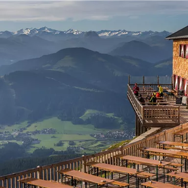 A mountain cabin with a large terrace and wooden chairs offers a stunning view of the surrounding mountains. The landscape is green with varying elevations and snow-capped peaks in the background.