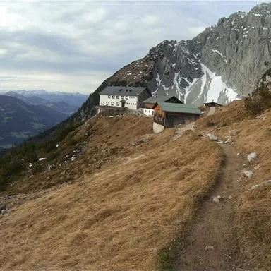 A picturesque mountain landscape with a hiking trail and historic buildings. In the background, majestic mountains and a cloudy sky can be seen.