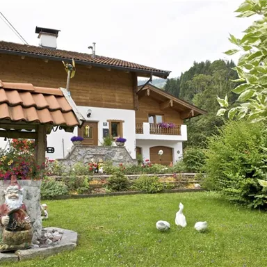 Ein traditionelles alpenländisches Haus mit einem aus Holz und Stein gebauten Brunnen. Im Vordergrund sind der Rasen und einige dekorative Figuren zu sehen.