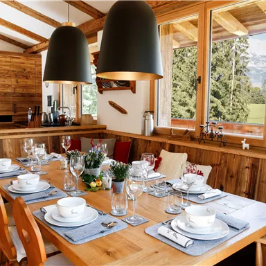 Ein einladender Essbereich aus Holz mit einem großen Tisch, der schön gedeckt ist. Große Fenster bieten einen Blick auf die grüne Landschaft und die Berge.