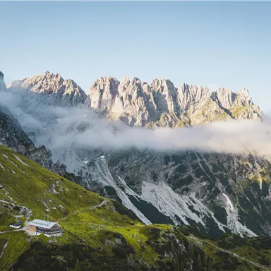 An impressive mountain landscape with majestic rocks and gentle hills. In the foreground, a small cabin is visible, surrounded by lush greenery and mist.