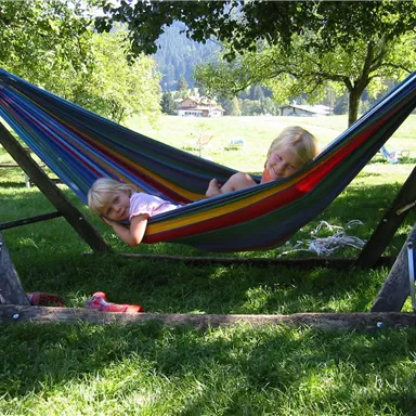 Two children are lying in a colorful hammock under a tree. The surroundings are green and sunny, with a view of a meadow in the background.
