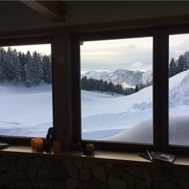 Ein Winterblick durch ein großes Fenster auf verschneite Berge und Wälder. Die Landschaft ist ruhig und malerisch mit einer sanften Schneedecke.