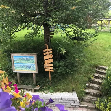 Ein malerischer Garten mit einer großen grünen Wiese und einem Baum. Im Vordergrund steht ein Holzschild und eine bunte Blumenrabatte.