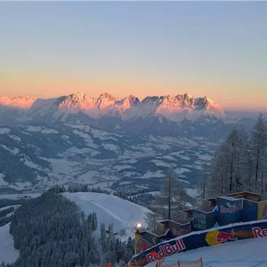 Eine winterliche Berglandschaft mit schneebedeckten Gipfeln und einem sanften Sonnenuntergang. Im Vordergrund ist eine skipiste zu sehen, die von einem Werbebanner gesäumt ist.