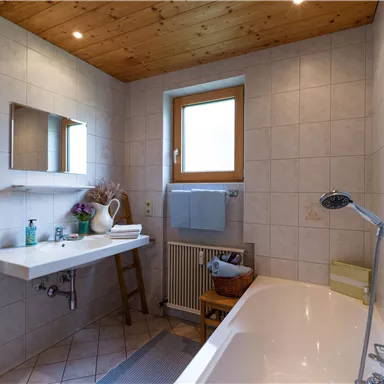 Ein modernes Badezimmer mit einer Badewanne und einem Waschbecken. Das Zimmer hat helle Fliesen und Holzdetails an der Decke.